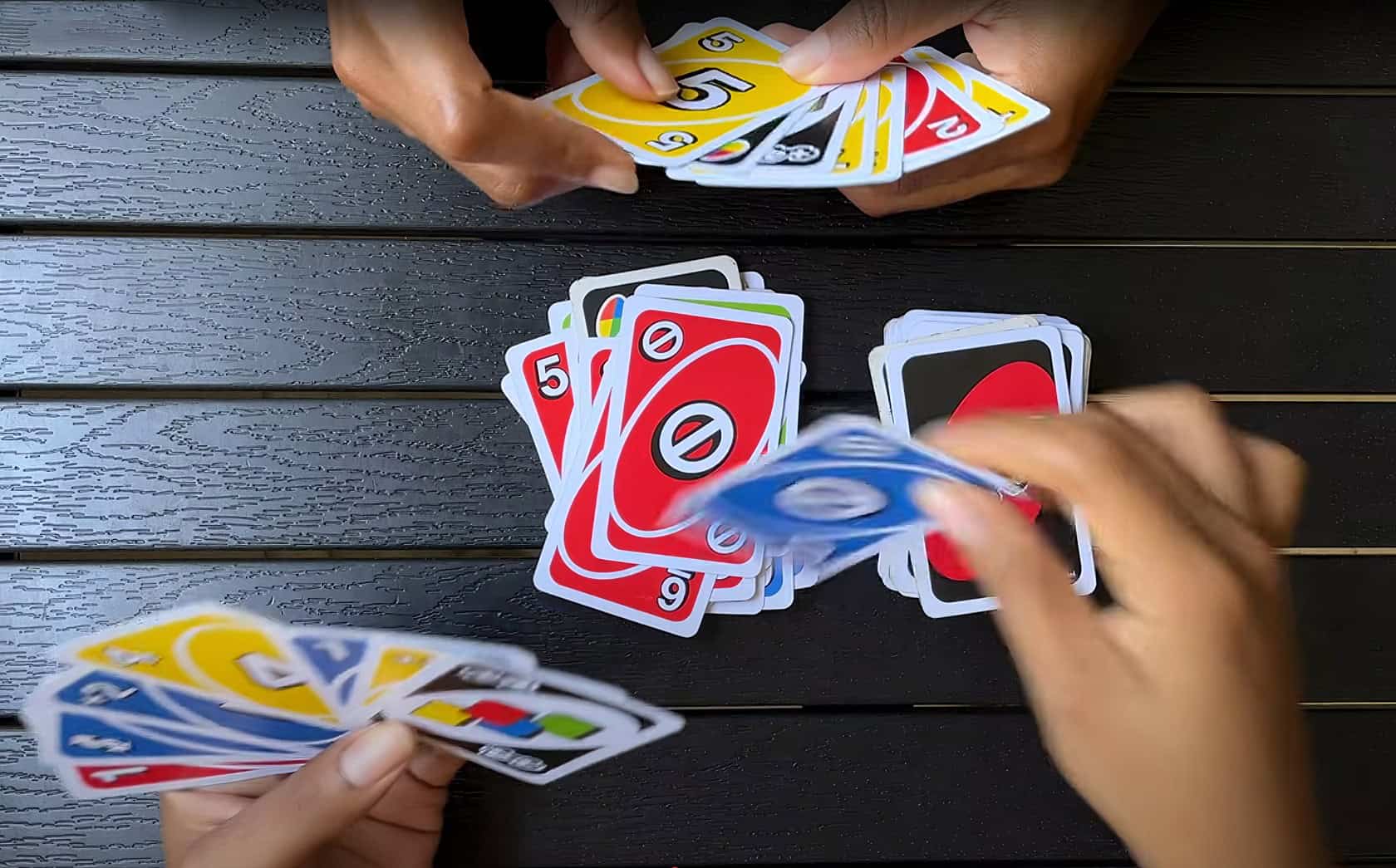 A pair of adult hands reaching for a deck of UNO cards on a wooden table in a cozy living room. What Does The Shuffle Hands Card Mean In UNO 2