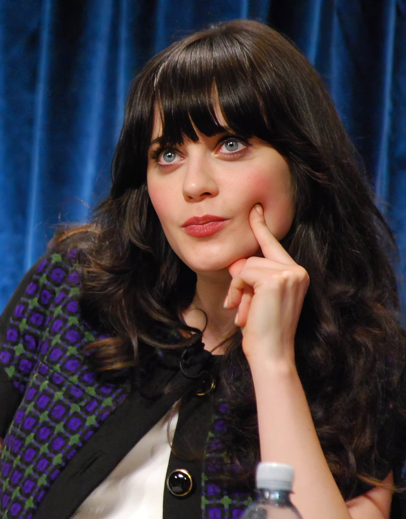 A woman in her 30s sits on a plush chair, playing a ukulele in a warmly lit, eclectic room. Zooey Deschanel