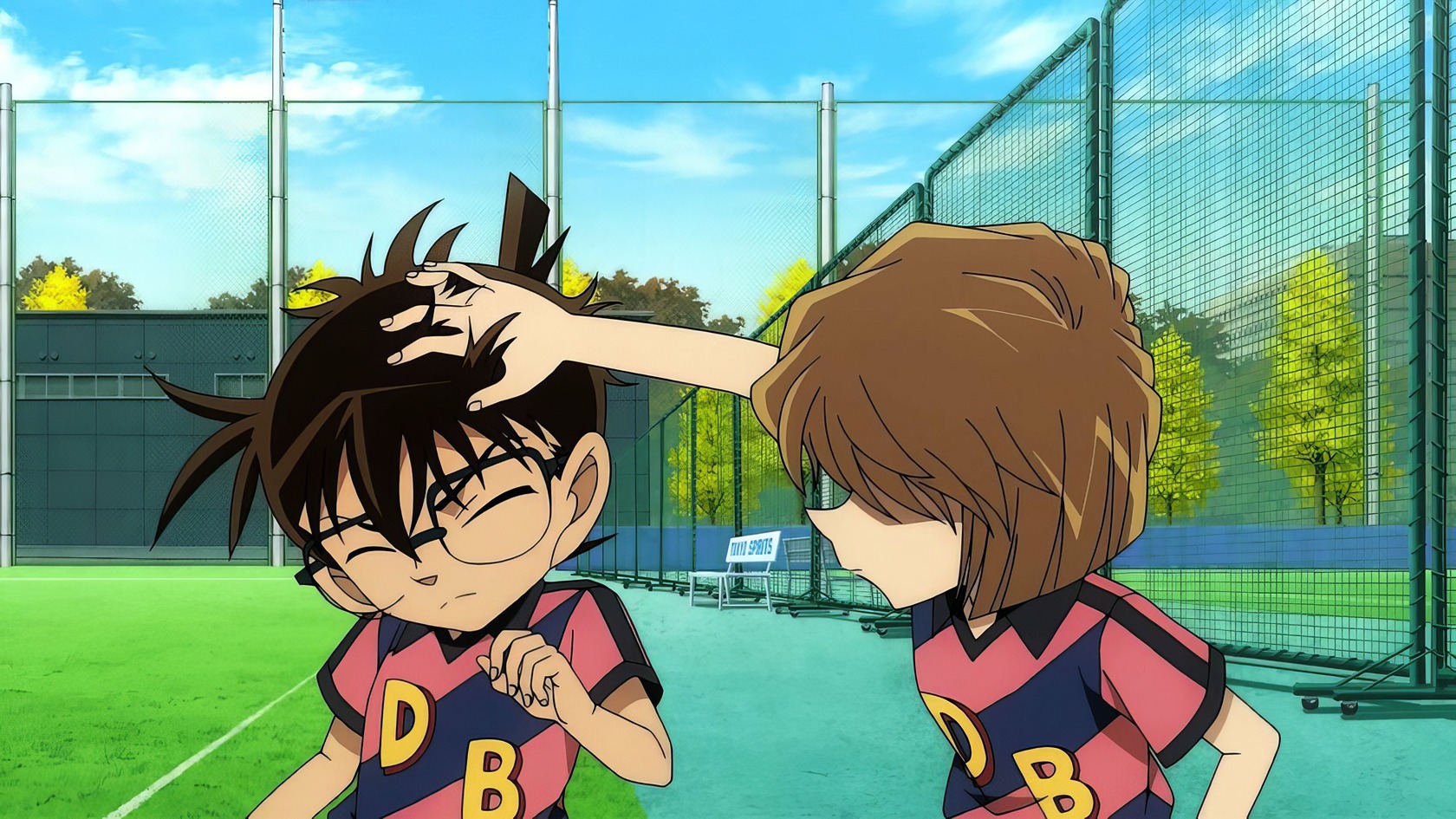 Anime About Soccer: 10 Winning Picks to Watch Now 7 Spiky-haired boy with glasses getting a head pat from girl with shoulder-length hair on an outdoor tennis court. Both are wearing sports jerseys with the letters "D" and "B" for team. Bright sky and lush trees in background.