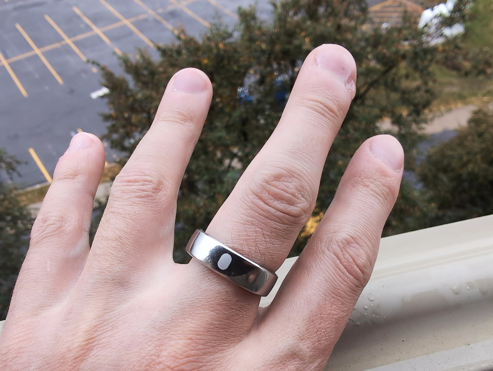 RingConn Gen 2 Air Review: 10 Reasons to Buy or Skip in 2025 3 A silver ring with a black dot on a person's finger, close-up of hand outdoors with a tree and parking lot in background.
