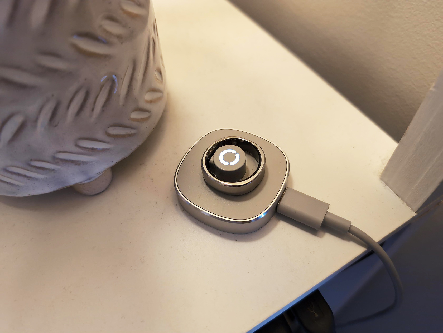 RingConn Gen 2 Air Review: 10 Reasons to Buy or Skip in 2025 6 Wireless charging pad connected to a device on a white surface, next to a decorative ceramic vase with a textured leaf pattern, in a modern home setting.