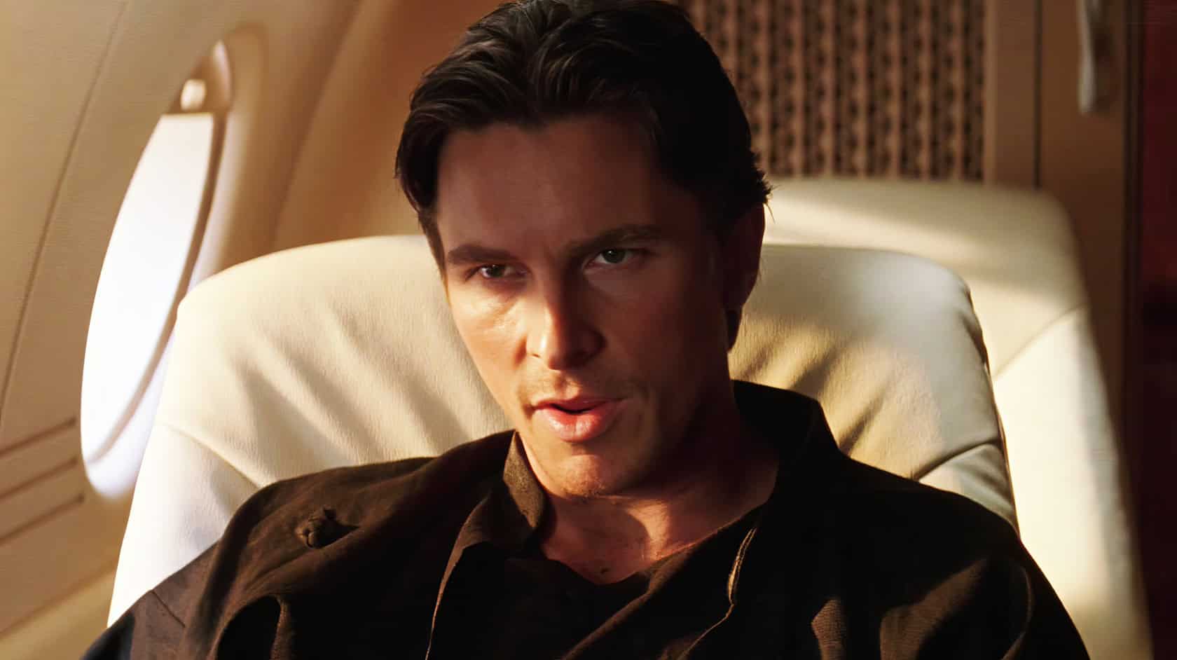 A man with dark hair and intense expression sitting in an airplane seat with beige leather upholstery.