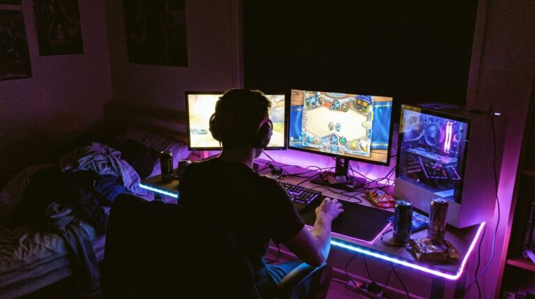 A person gaming on a multi-monitor setup with RGB lighting in a dark room, surrounded by gaming accessories and snacks, showcasing an immersive gaming environment for esports and gaming enthusiasts.