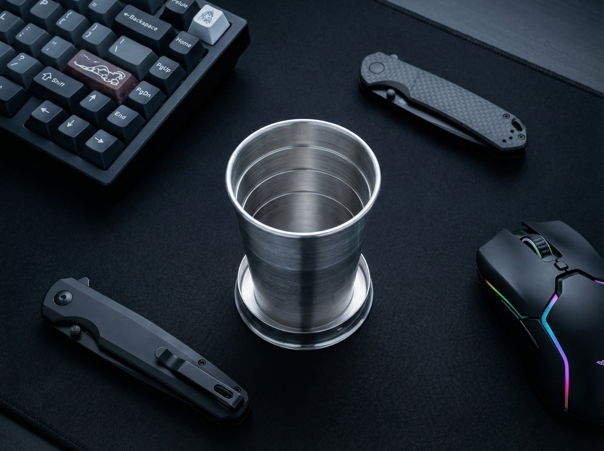 Review: Mizu Collapsible Shot Glass for Emergency Drinking 3 High-quality gaming mouse, folding knife, mechanical keyboard with custom keys, and a stainless steel cup on a black workspace emphasizing gaming and tech accessories.