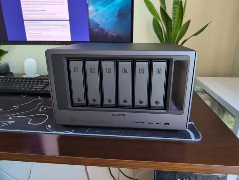 An image of the UGREEN iDX NAS device with six drive bays labeled 01 to 06, placed on a wooden desk with a computer monitor and a green plant in the background.