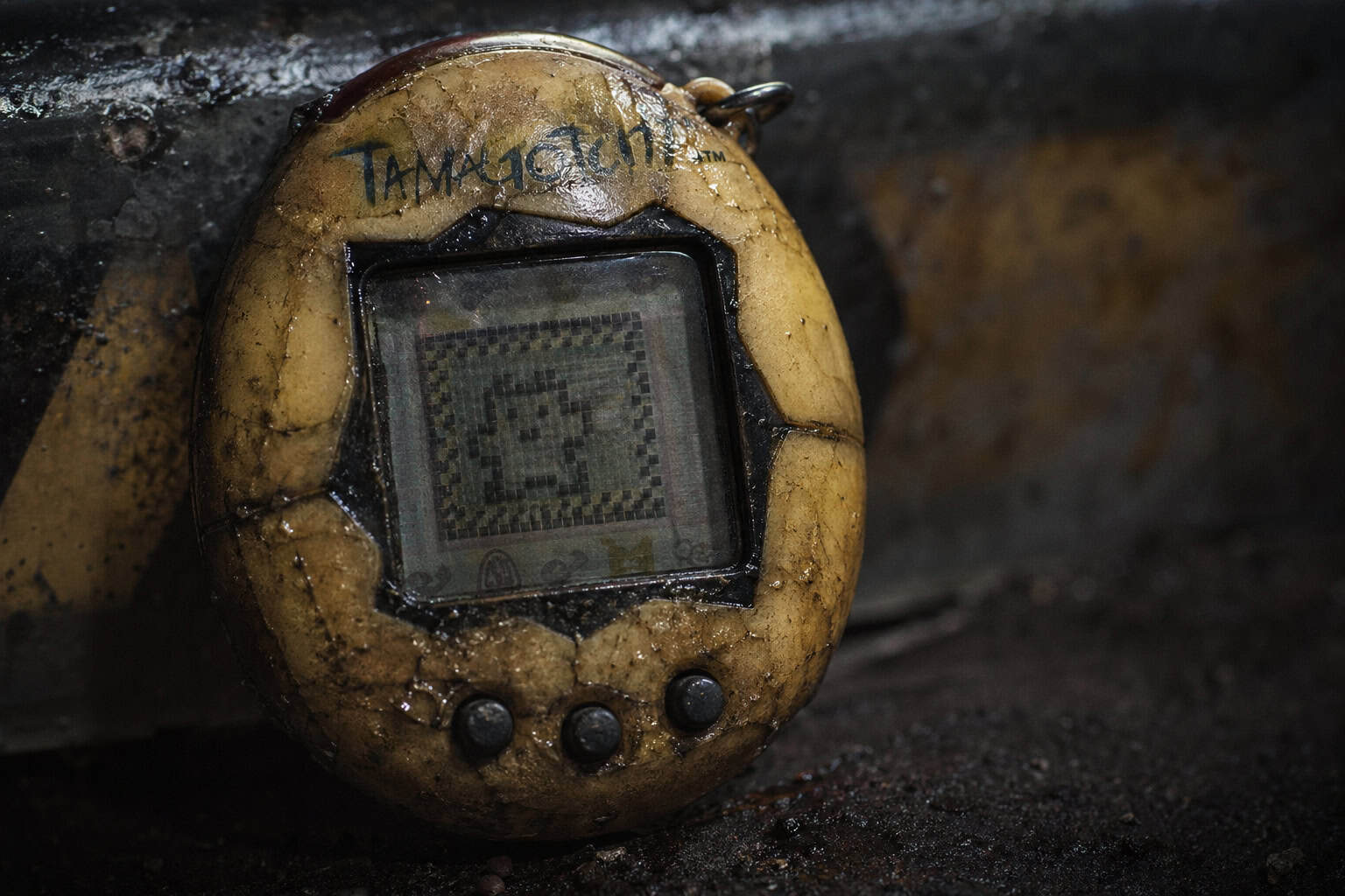 A digital pet device resembling a Tamagotchi, covered in dirt and grime, lying on a rough, dark surface. The device appears old and damaged, with visible scratches and dirt, emphasizing a rugged, aban.
