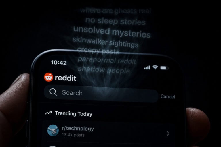 Close-up of a smartphone displaying Reddit app with trending posts, with ghostly text in the background mentioning ghosts, mysteries, and paranormal themes.