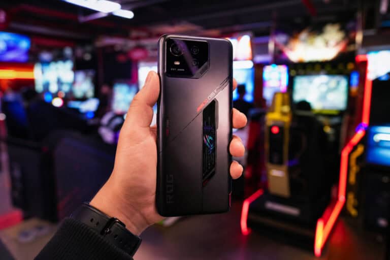 Close-up of a person holding an ASUS ROG Phone 6 gaming smartphone in a gaming arcade with colorful neon lights and arcade machines in the background.
