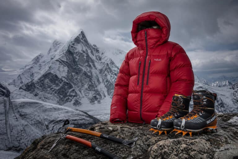Climber's red insulated jacket, ice axes, and crampons on rocky mountain terrain with snow-capped peaks in background.