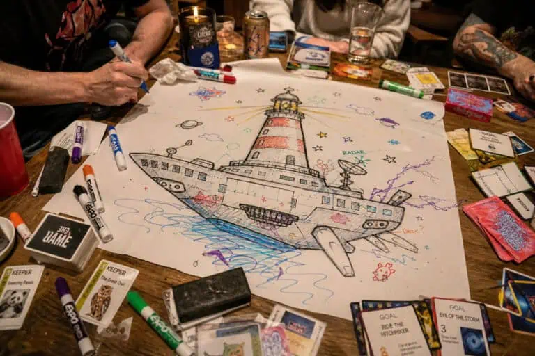 Group of people drawing a colorful lighthouse and ship on a large sheet of paper with markers and game cards around.