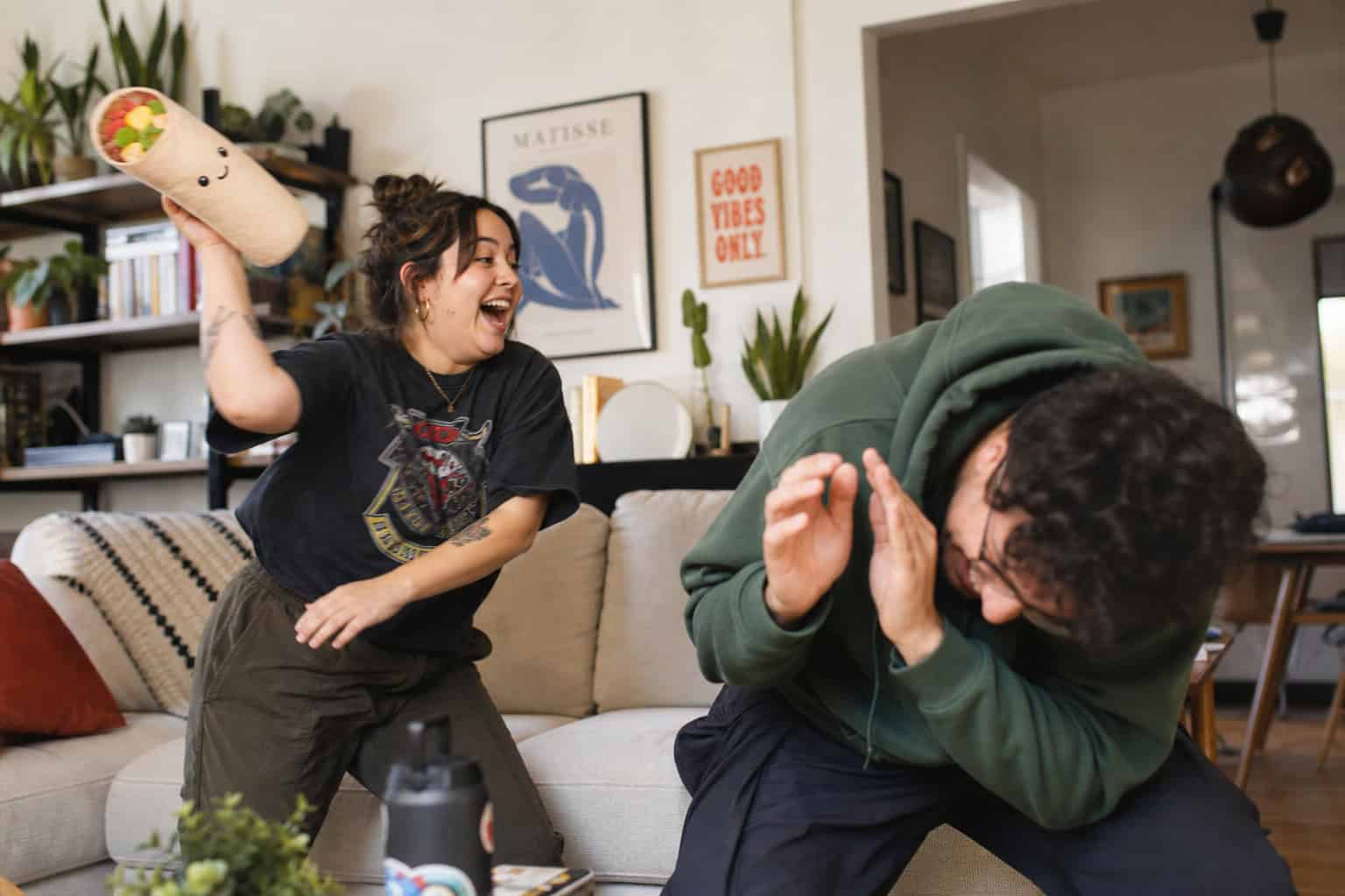 10 Free Online Party Games for Adults That Actually Work (Ranked) 5 A woman and a man are having fun in a cozy living room, with the woman playfully hitting the man with a plush toy, capturing a joyful moment.