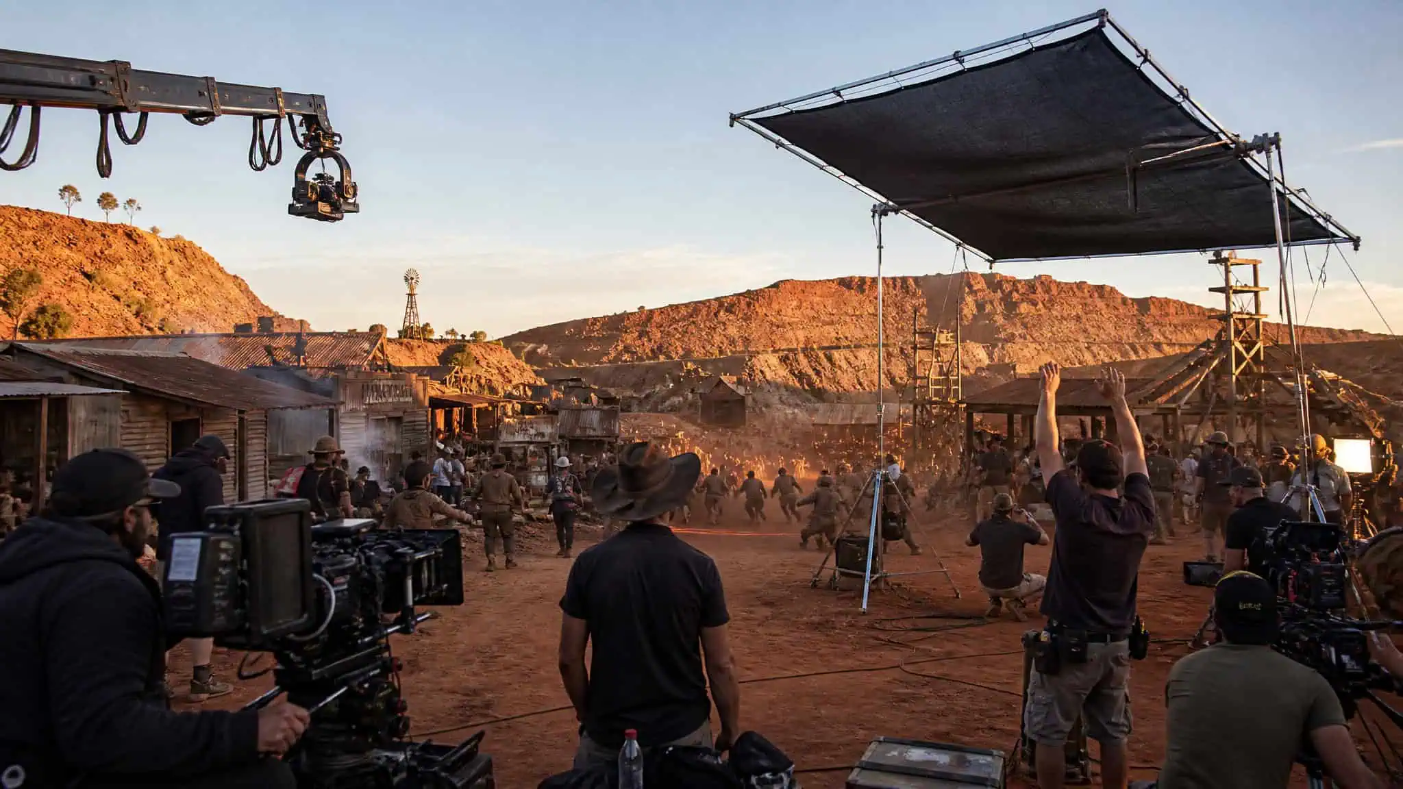 Film crew working on a Western movie set in a desert landscape with mountains in the background, using professional cameras and lighting equipment.