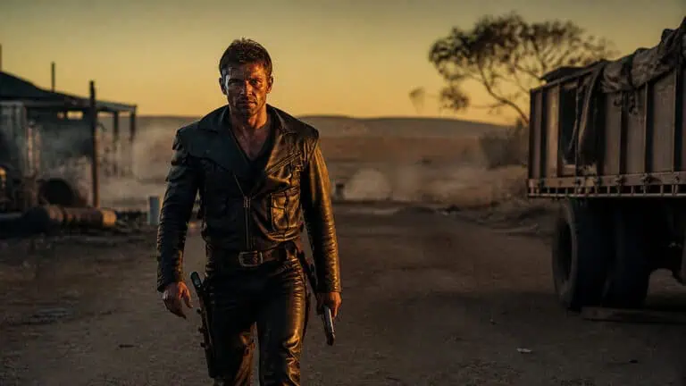 A rugged man in a black leather jacket holding a gun, walking through a dusty desert at sunset with trucks and sparse trees in the background.