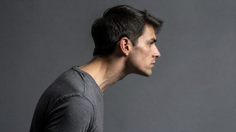 Side profile of a young man with dark hair and a serious expression, wearing a gray shirt against a plain gray background.