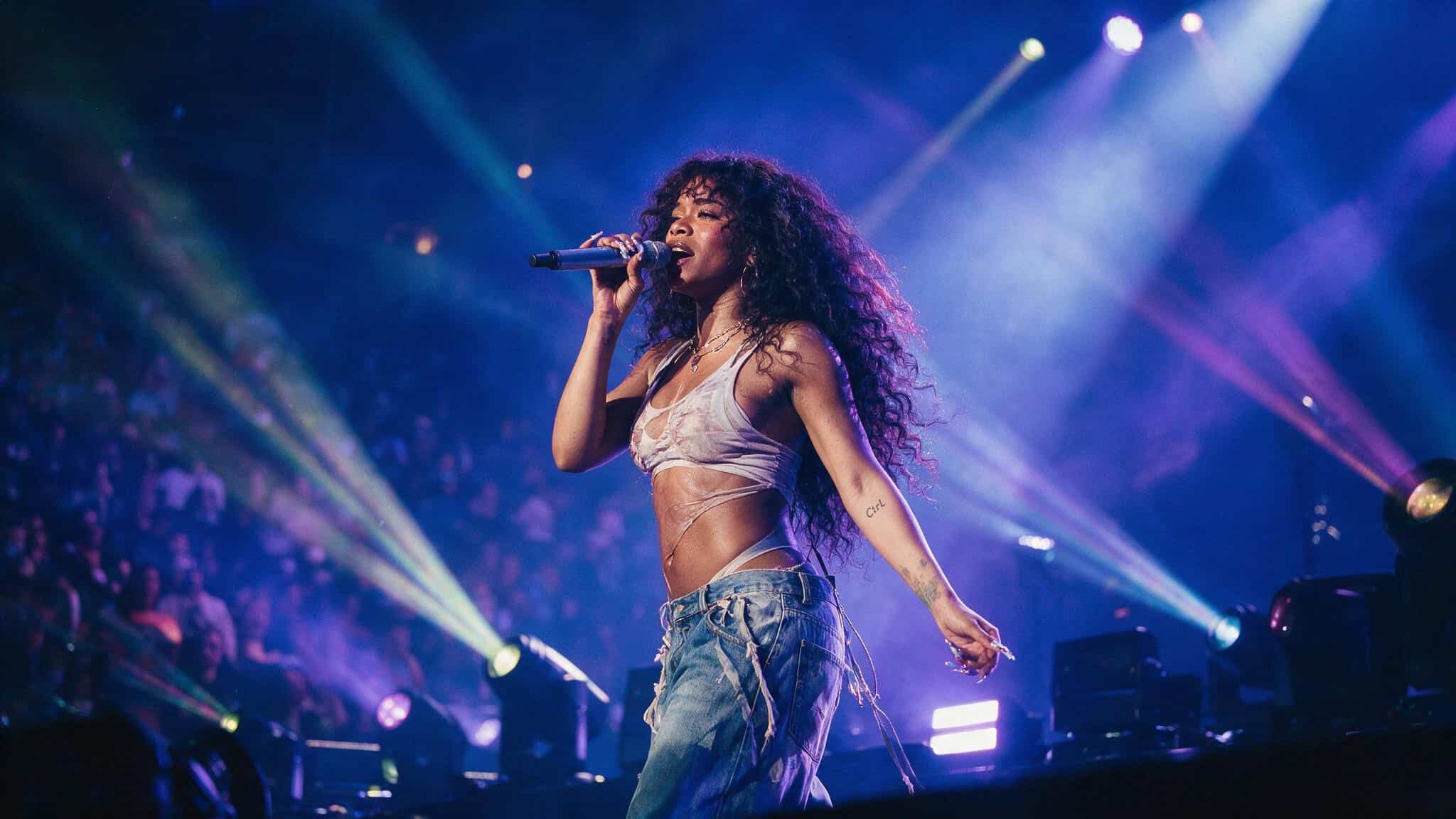 Female singer with curly hair performing live on stage with colorful lights and audience in background.