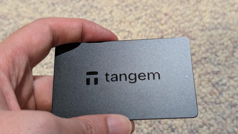 Close-up of a Tangem crypto wallet card held in hand on a sandy surface, showcasing its sleek design and branding.