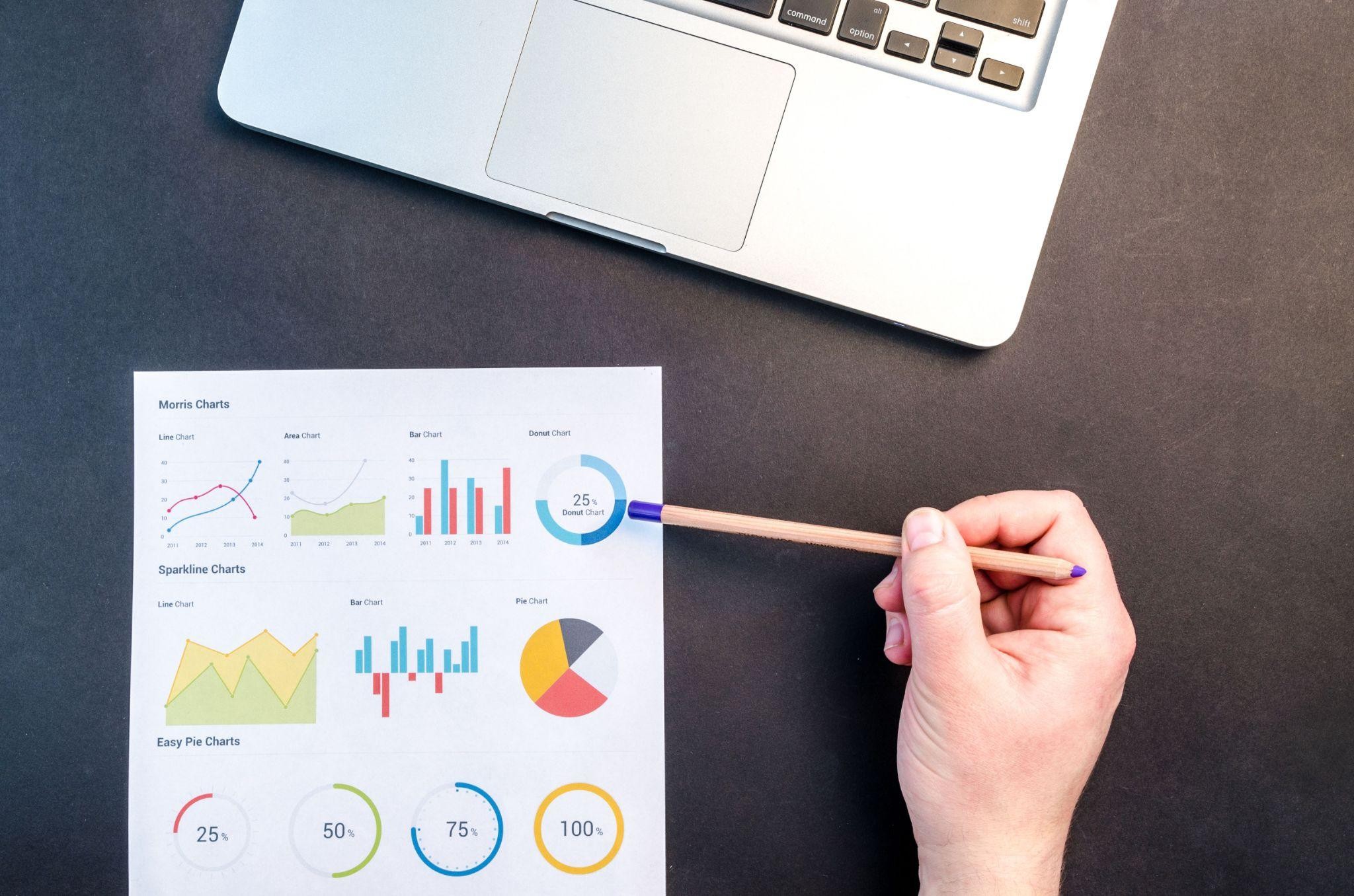 Person analyzing data charts and graphs with a pencil near a laptop on a desk.