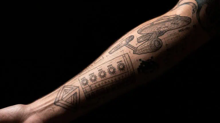 Close-up of a tattooed arm featuring tech-inspired designs including circuit boards, pixel art, and digital elements on a black background.