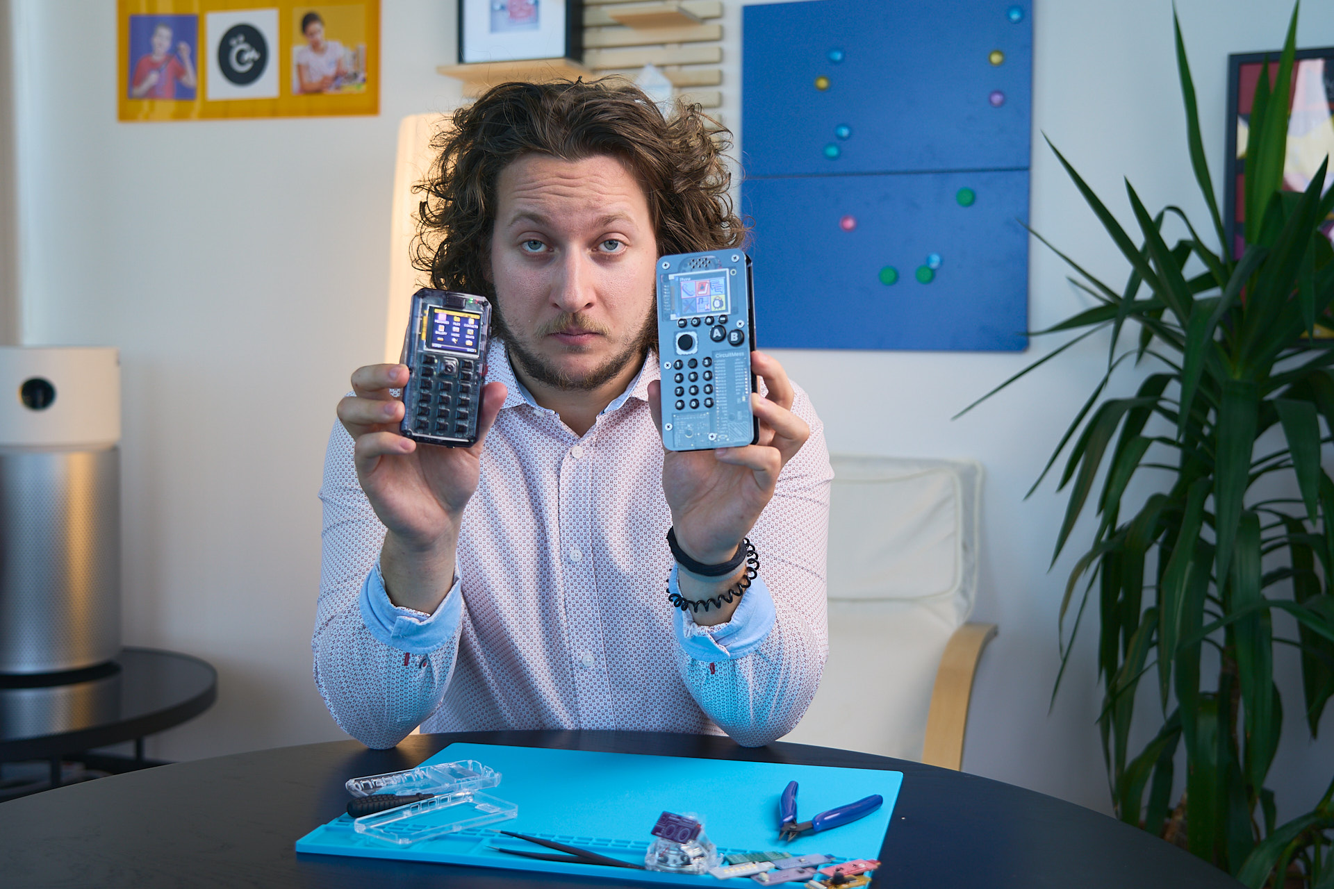 MAKERphone 2.0: Every Pledge Tier From $109 to $7,777, Decoded 3 Man with curly hair holding two remote controls, showing them to the camera in a modern room setting.