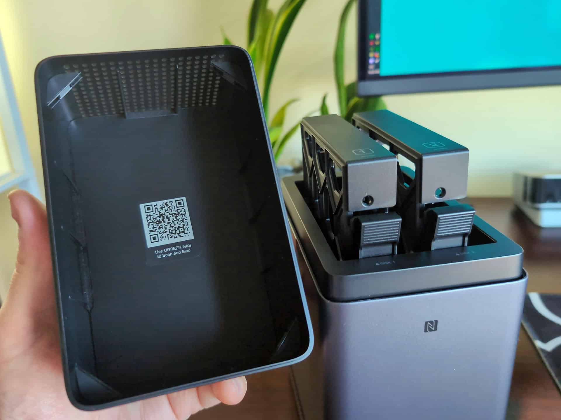 Ugreen DH2300 vs DXP2800 GT: Which Two-Bay NASync Should a First-Time Buyer Actually Get? 4 Close-up of Ugreen NAS docking station with two hard drives and a smartphone in the foreground, on a desk with a monitor and plant in the background.