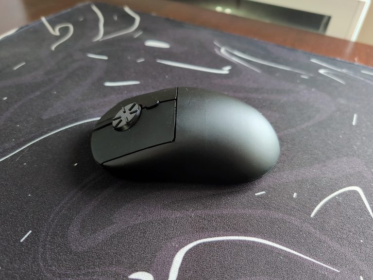 Wireless rotary mouse with a dial on top, placed on a black mouse pad with white markings, in a well-lit room.