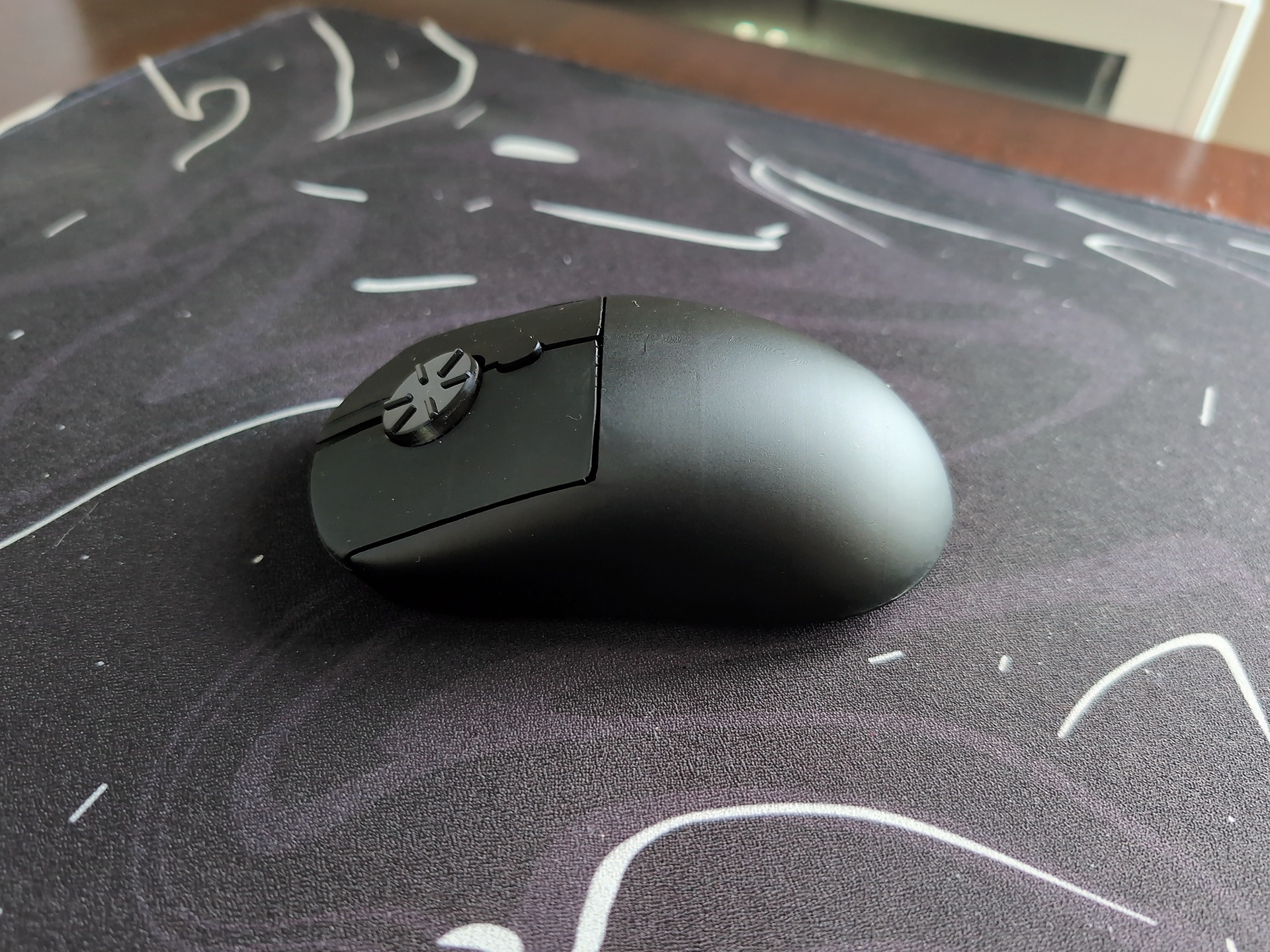 Wireless rotary mouse with a dial on top, placed on a black mouse pad with white markings, in a well-lit room.