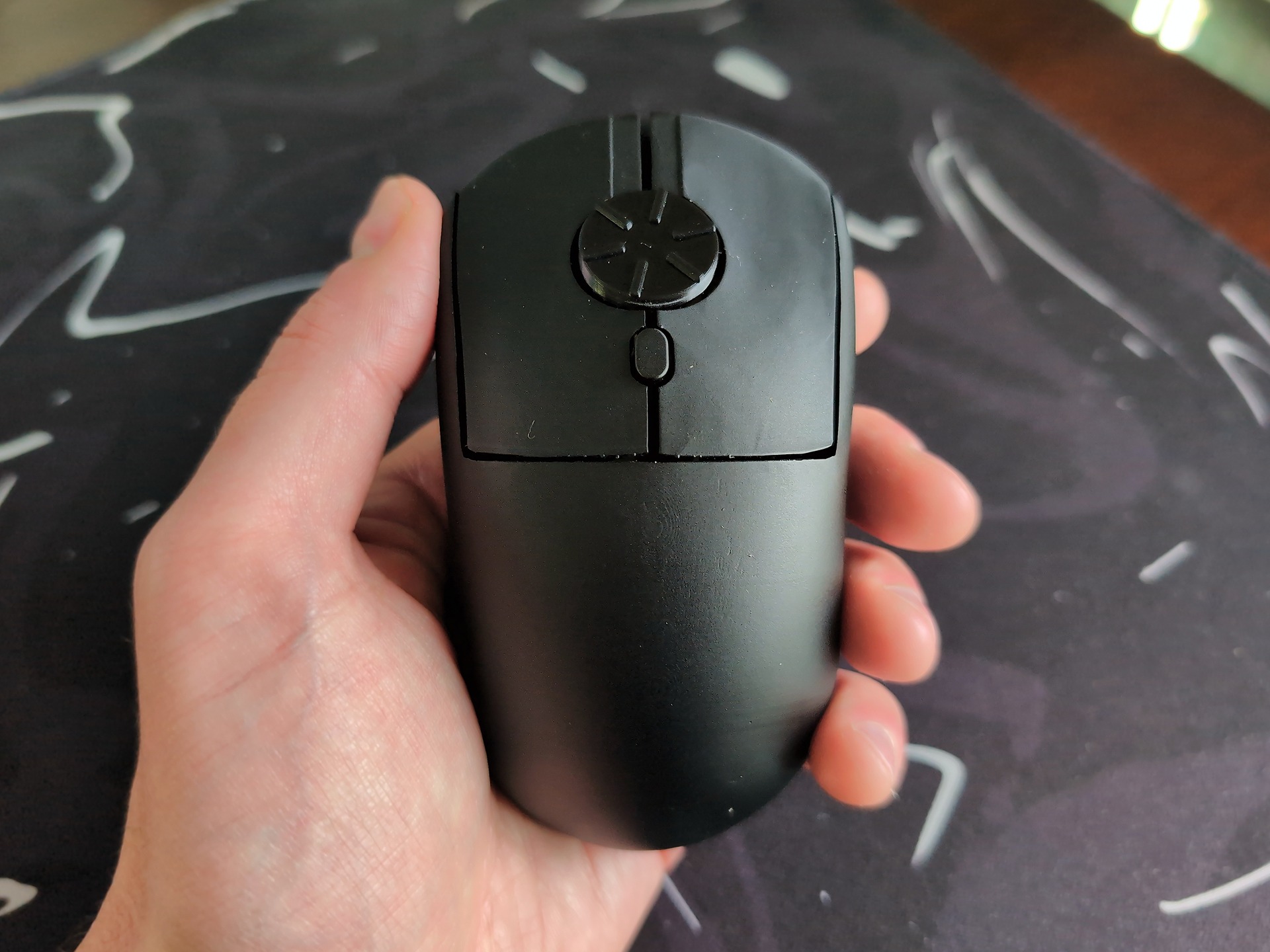 Rotary Mouse Review: 3 Weeks With the Dial That Replaced My Scroll Wheel 2 Close-up of a black rotary mouse with a dial on top, held in a person's hand, on a dark surface with white markings.